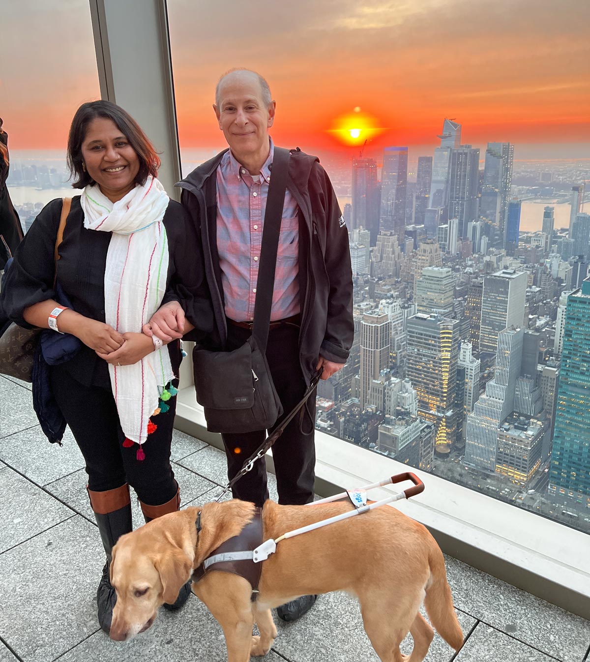Lakshmee Lachhman-Persad and Peter Slatin along with Peter's guide dog Inga at the Summit One Vanderbilt with the sun setting over the stunning backdrop of manhattan's skyline.