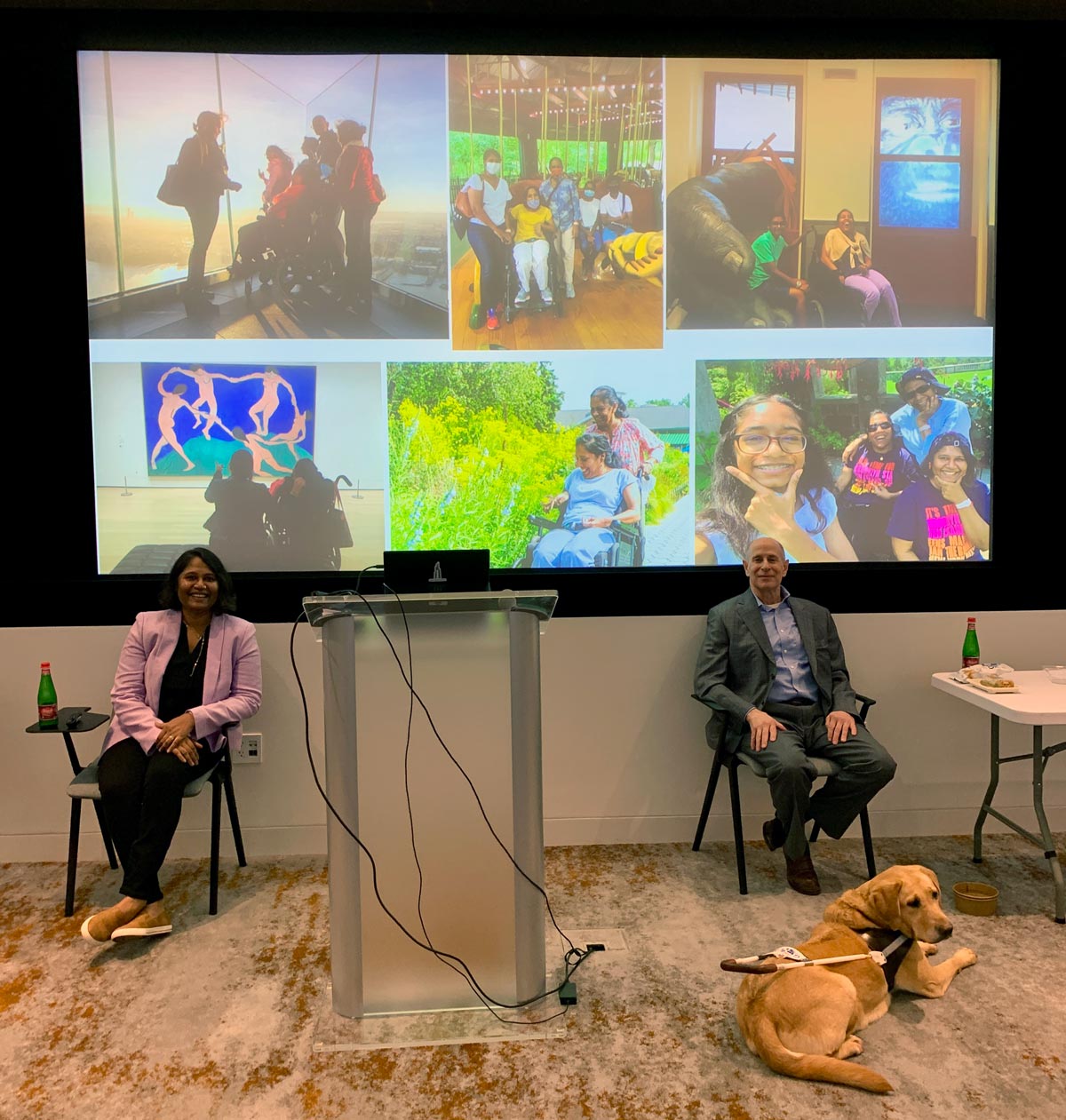 Lakshmee Lachhman-Persad and Peter Slatin along with Peter's guide dog Inga at the Elements of Service training at the Summit Observatory at One Vanderbilt, seated in front of the projection featuring various photographs.
