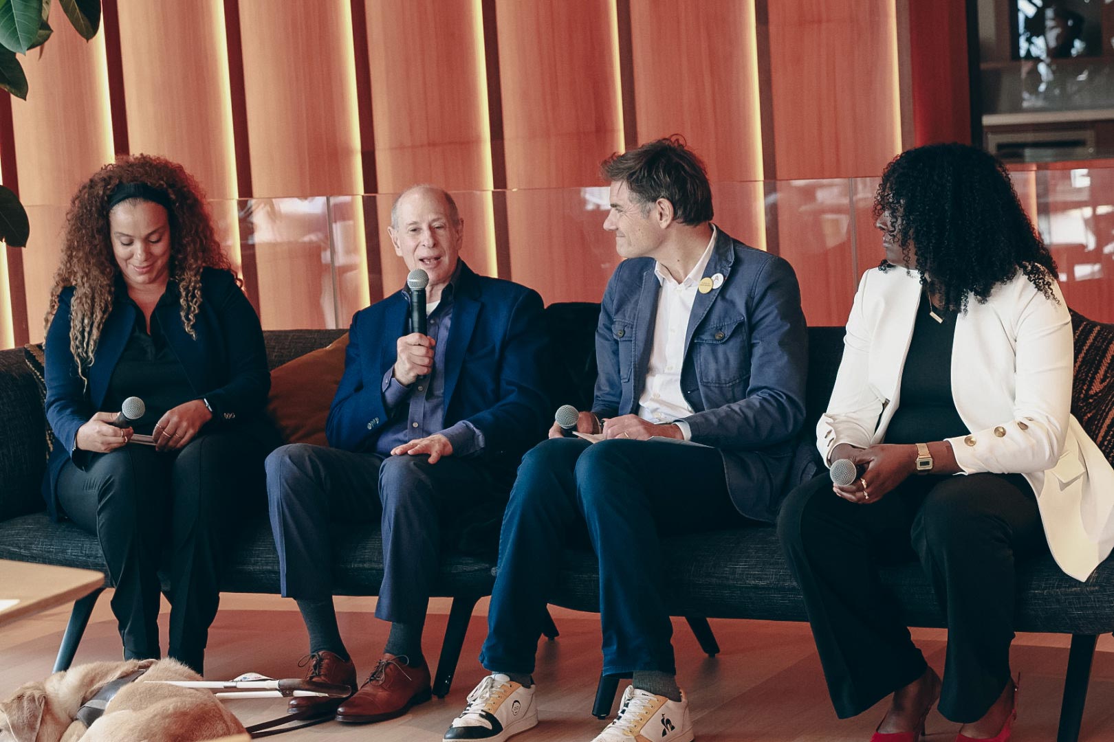 From Left to right Claribel, Peter, Yann, Alyssia seated on a black couch. Peter is holding a microphone speaking during the panel, there is a dog laying on the floor in front of peter.