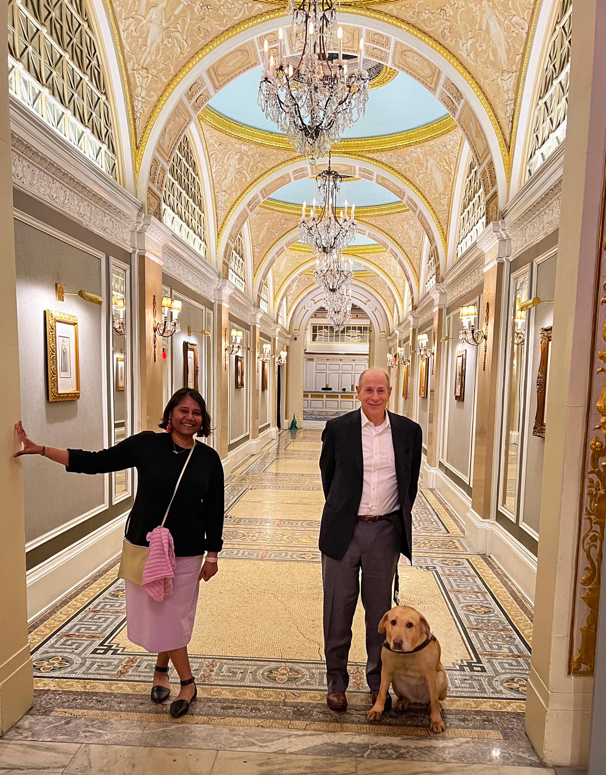 Lakshmee Lachhman-Persad and Peter Slatin with his guide dog Inga standing in the lobby of the historic Fairmont hotel in a long, arched, opulent hallway featuring grand chandeliers, artwork on the walls and a mosaic floor.