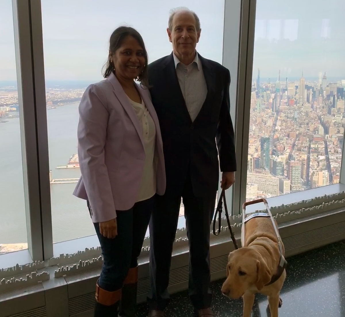Lakshmee Lachhman-Persad (left), Peter Slatin (right) and Peter's guide dog Inga at the One World Observatory, Manhattan view in the background