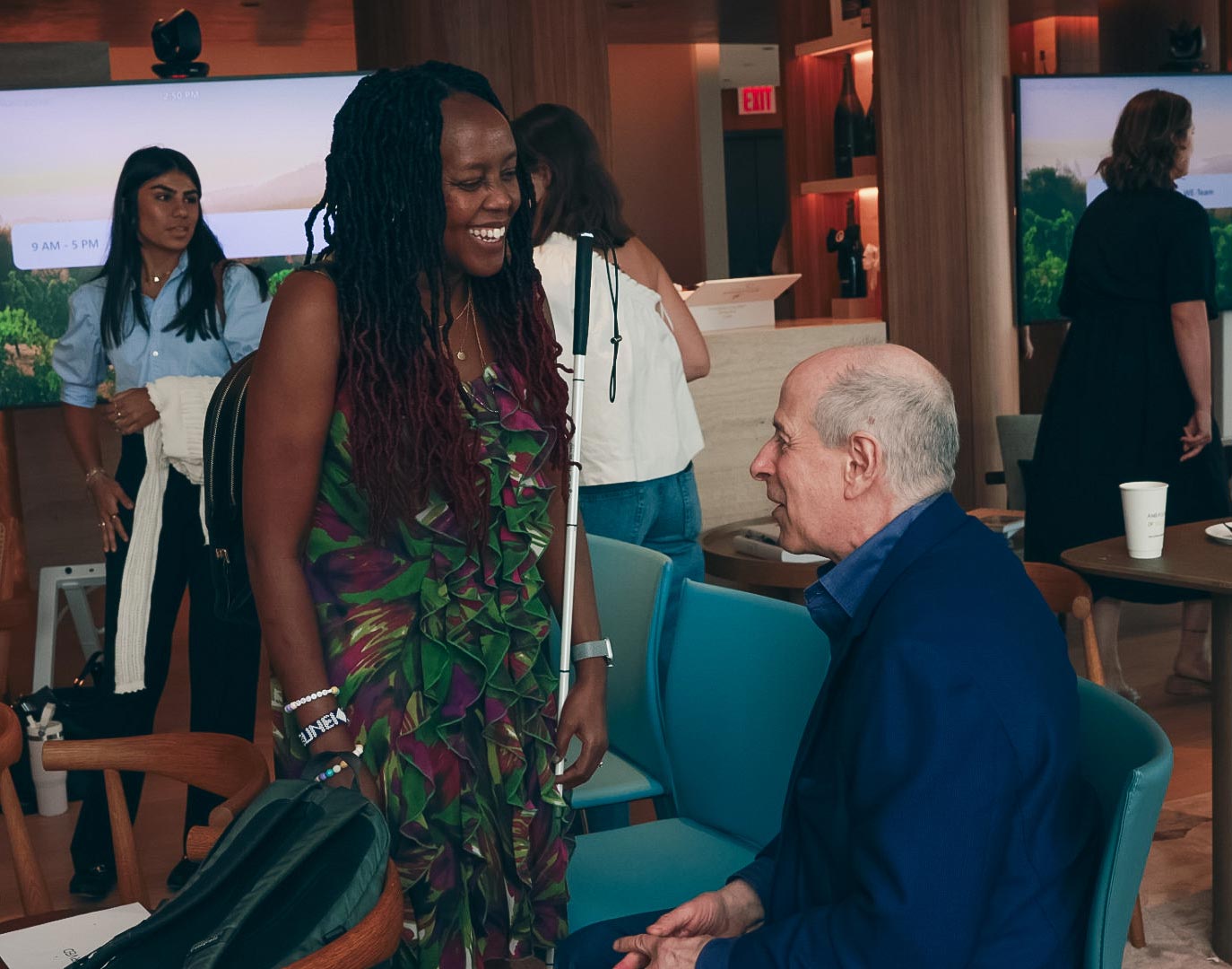 Loice holds up a white cane while chatting with Peter who is seated in a teal chair inside a modern lounge. Other people mingle in the background near large screens and tables with cups.
