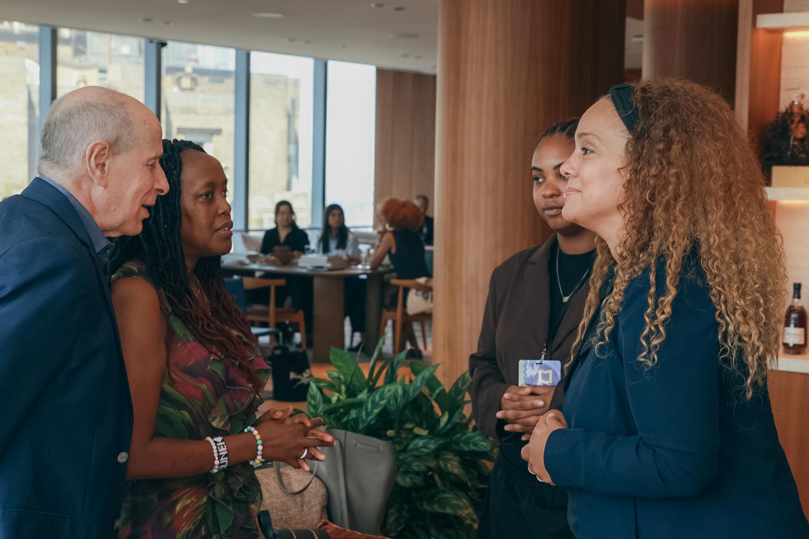 Peter and Loice standing on the left side across from Arielle and Claribel, the group is talking to eachother before the event in the library area. Arielle introduced Claribel to Peter and Loice. There are a few people pictured in the back seated at a table.